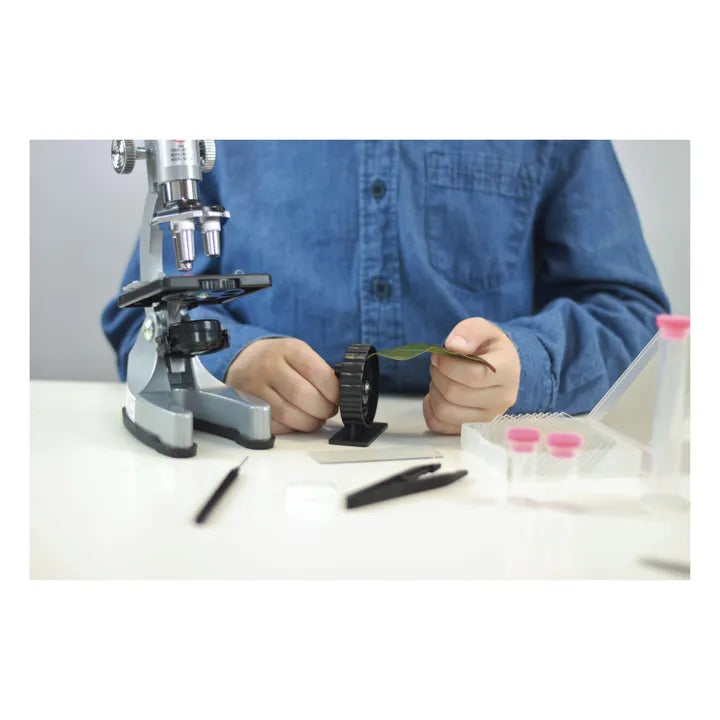 Mãos de uma criança a manusear uma folha e uma lupa de mão, com o microscópio Buki e outros acessórios em segundo plano.