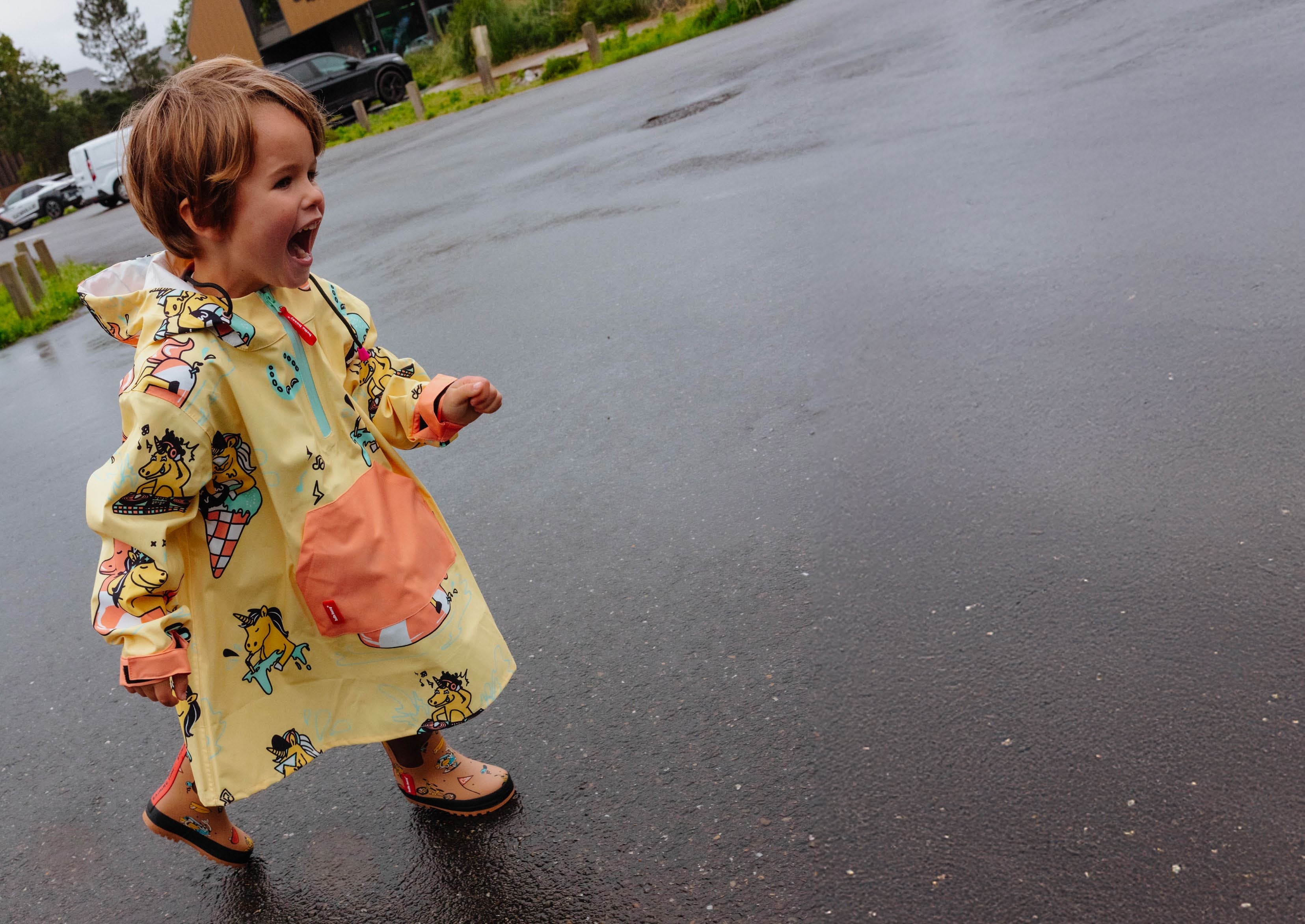 Criança pequena a correr e sorrir ao ar livre, vestindo o poncho Hello Hossy Unicorn amarelo com padrão de unicórnios em dia de chuva.