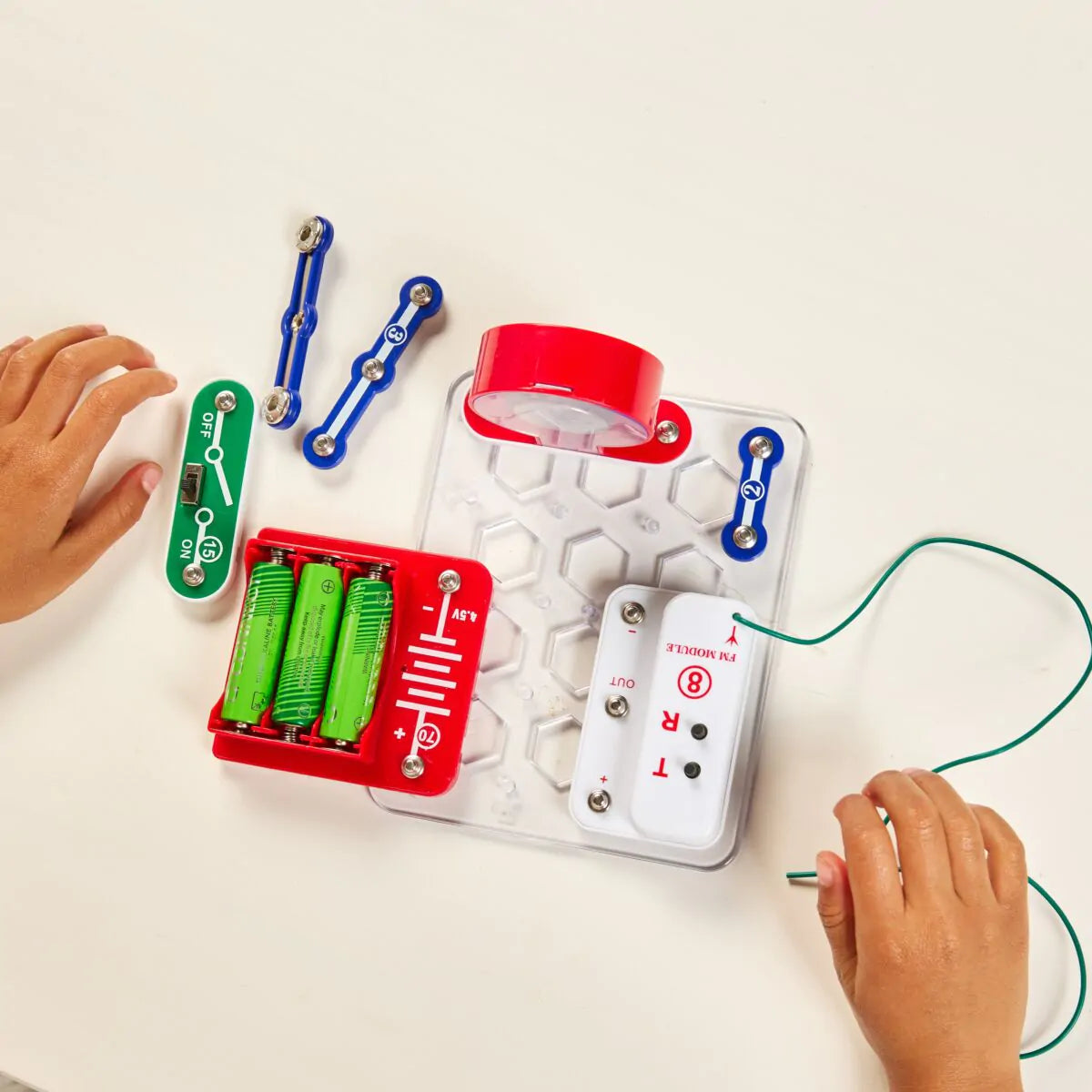 Mãos de criança a montar o kit Miniland Electrofun: Tune In Radio, com as peças modulares, o compartimento de pilhas e o módulo FM visíveis sobre uma mesa branca.