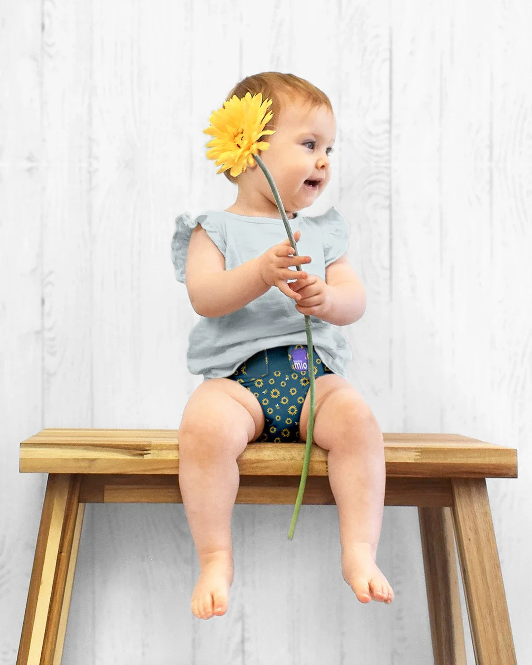 Bebé sentado num banco de madeira, a usar uma fralda reutilizável Bambino Mio miosolo classic com padrão de girassóis, segurando uma flor amarela.