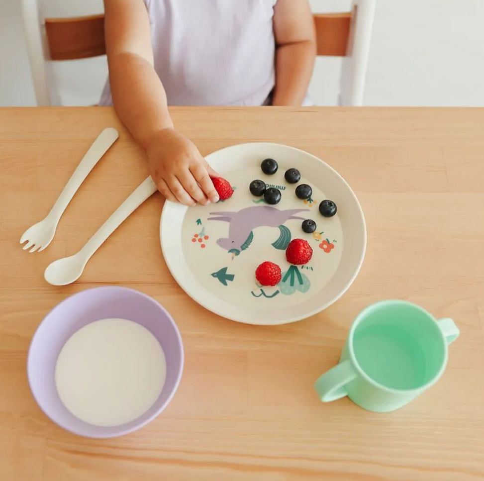 Criança a comer frutas do prato Miniland Dream com unicórnio, tigela, copo e talheres na mesa de madeira.