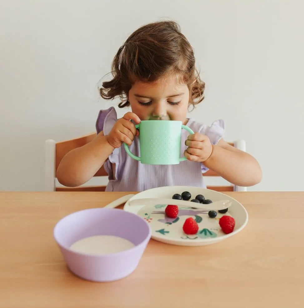 Criança a beber do copo de aprendizagem Miniland Dream verde com alças ergonómicas, na mesa.
