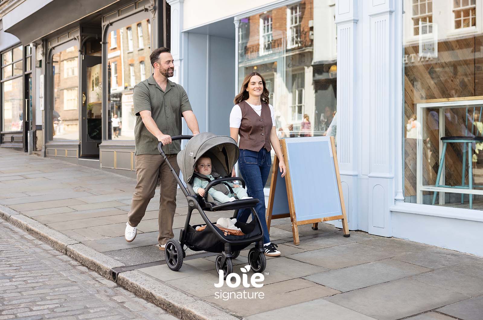Casal a passear com o carrinho Joie Finiti Signature e bebé no assento virado para a frente, em rua comercial.