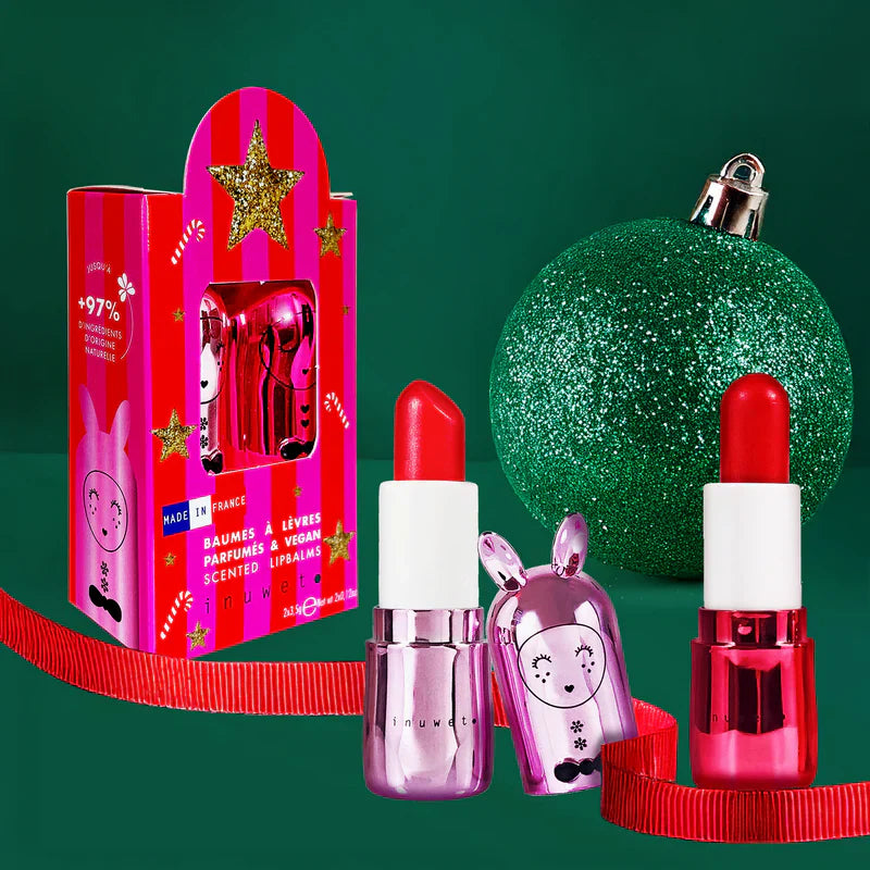 Conjunto de 2 bálsamos labiais Inuwet e a sua embalagem festiva, com um enfeite de Natal verde e uma fita vermelha, sobre um fundo verde escuro.