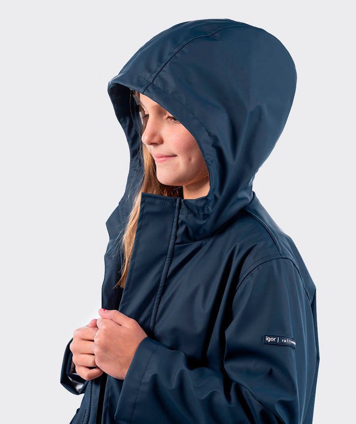 Criança com o casaco impermeável Igor azul-marinho e capuz, vista de perfil, sorridente.