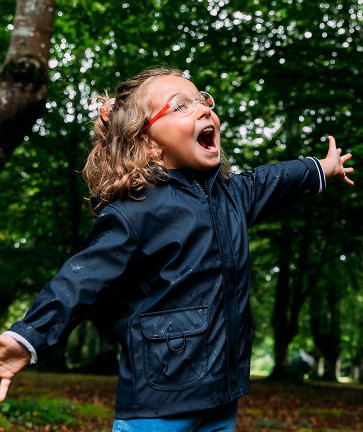 Criança feliz a usar o casaco impermeável Igor azul-marinho num ambiente natural, com os braços abertos.