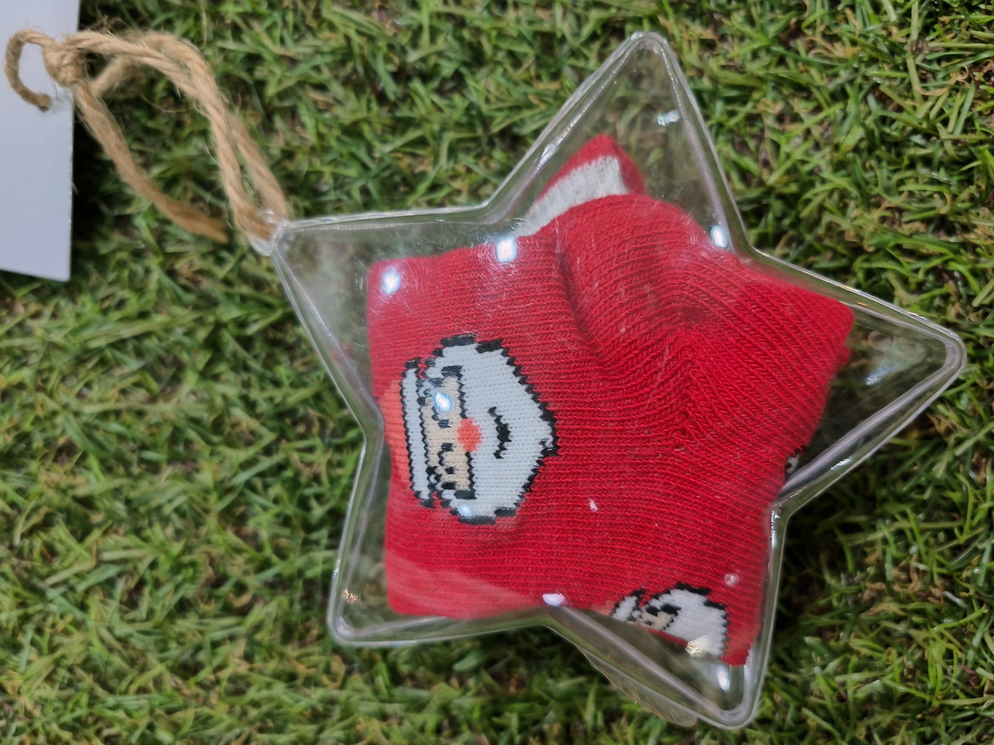Meias de bebé Ciocca vermelhas com padrão de Pai Natal, dobradas dentro de uma caixinha transparente em forma de estrela, sobre um fundo de relva artificial.
