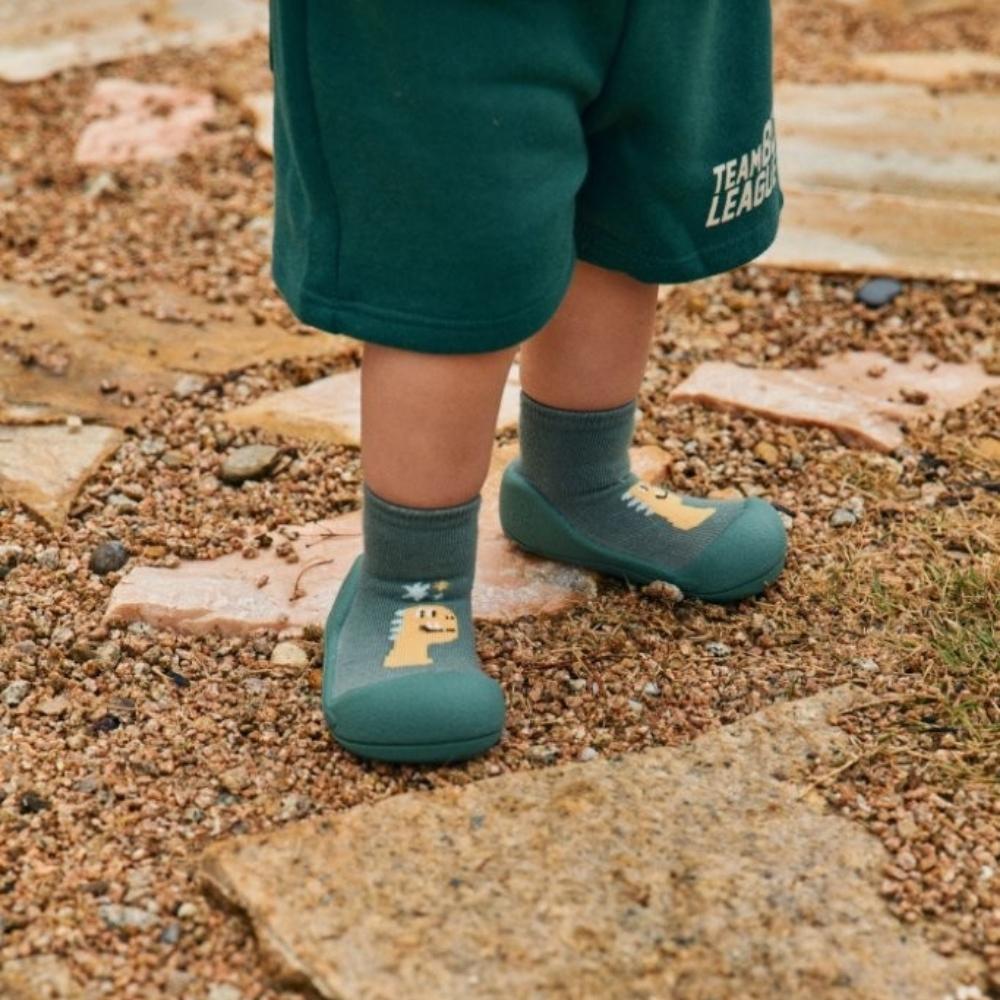 Bebé a usar sapatos Attipas DINO GREEN, com calções verdes, a dar passos numa área exterior com pedras e terra, mostrando a flexibilidade do calçado.