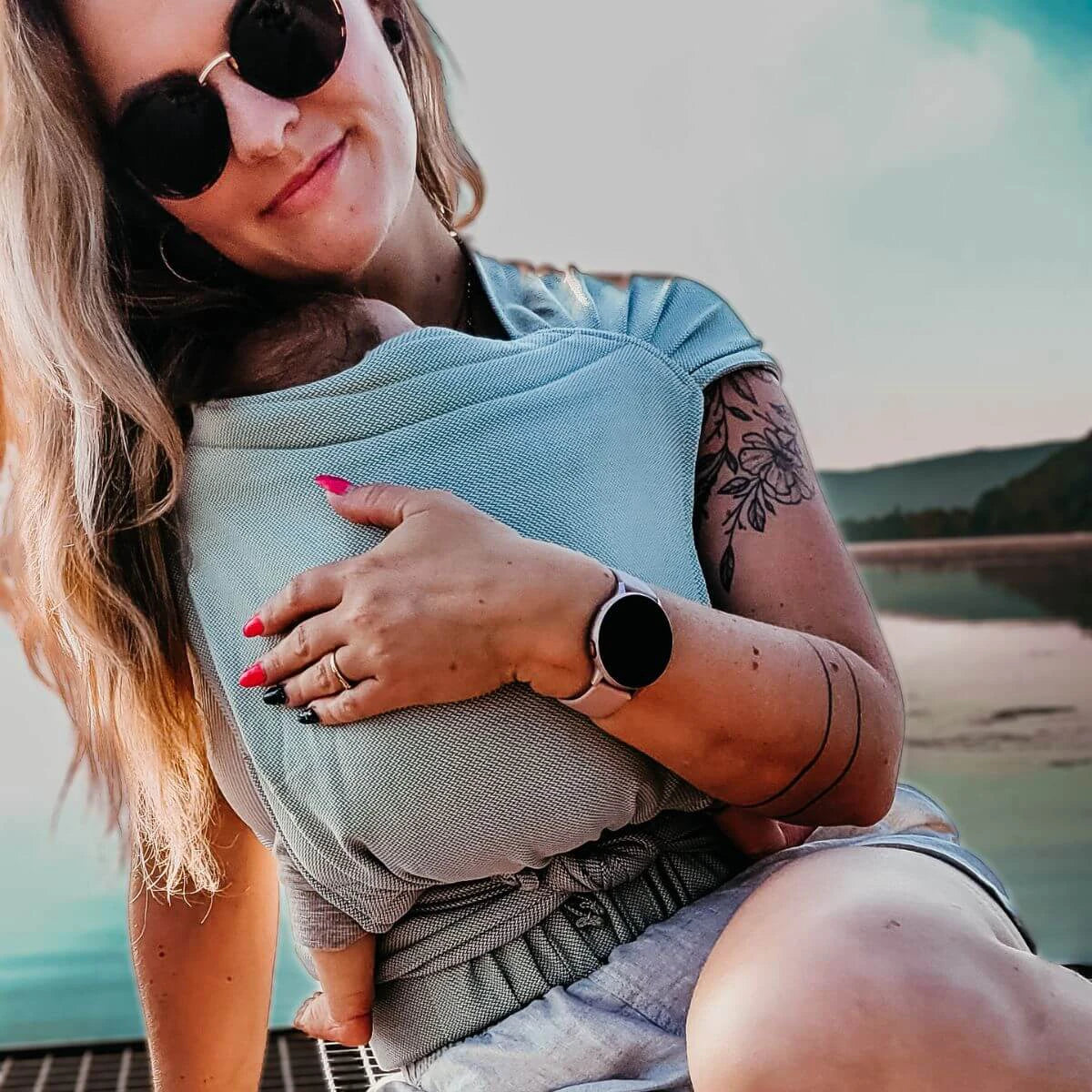 Mulher loira a usar o porta-bebé HOPPEDIZ® cinza, segurando um bebé pequeno e sorrindo.