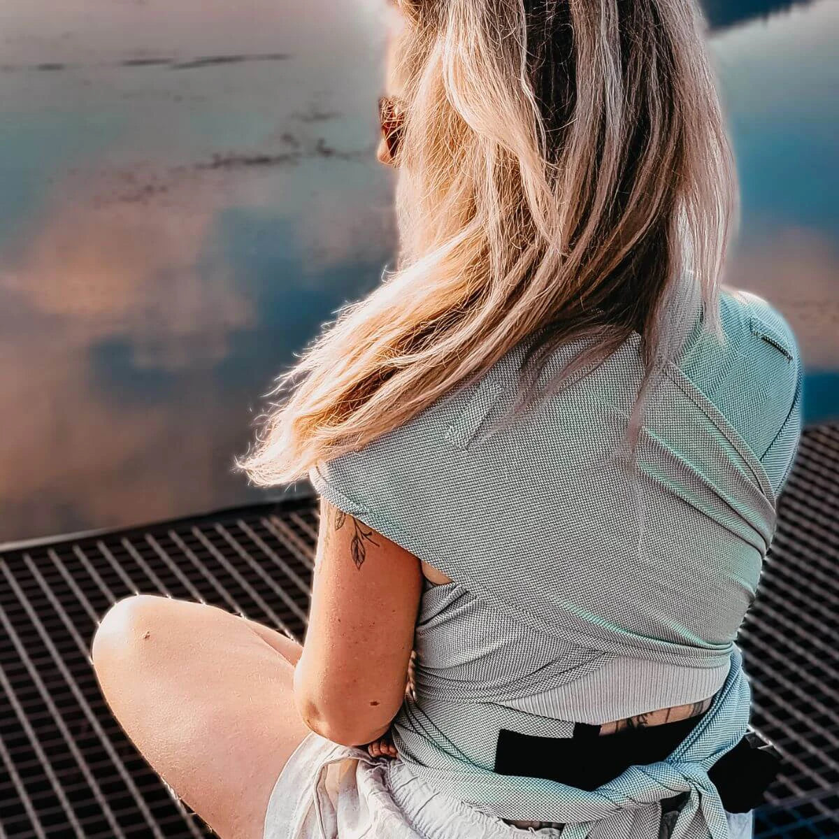 Vista traseira da mulher a usar o porta-bebé HOPPEDIZ®, destacando a amarração e o capuz enrolado.