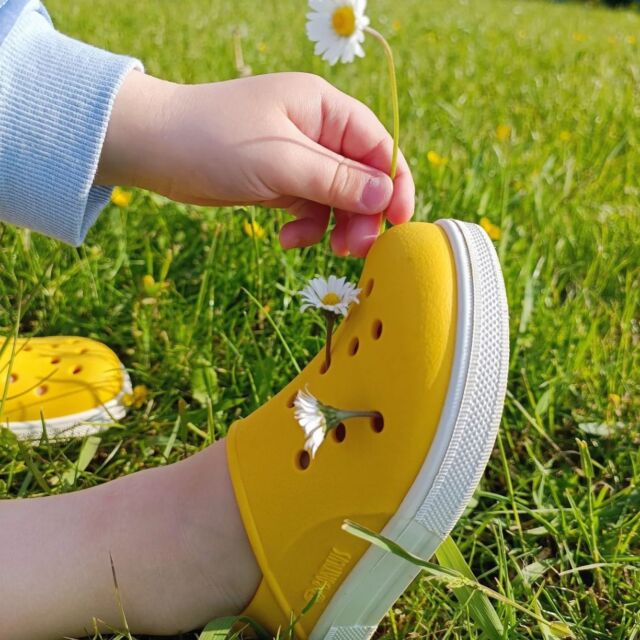 Criança a usar sandália Boatilus Cloggy Amarelo no pé, a brincar na relva com uma flor, destacando o uso exterior.