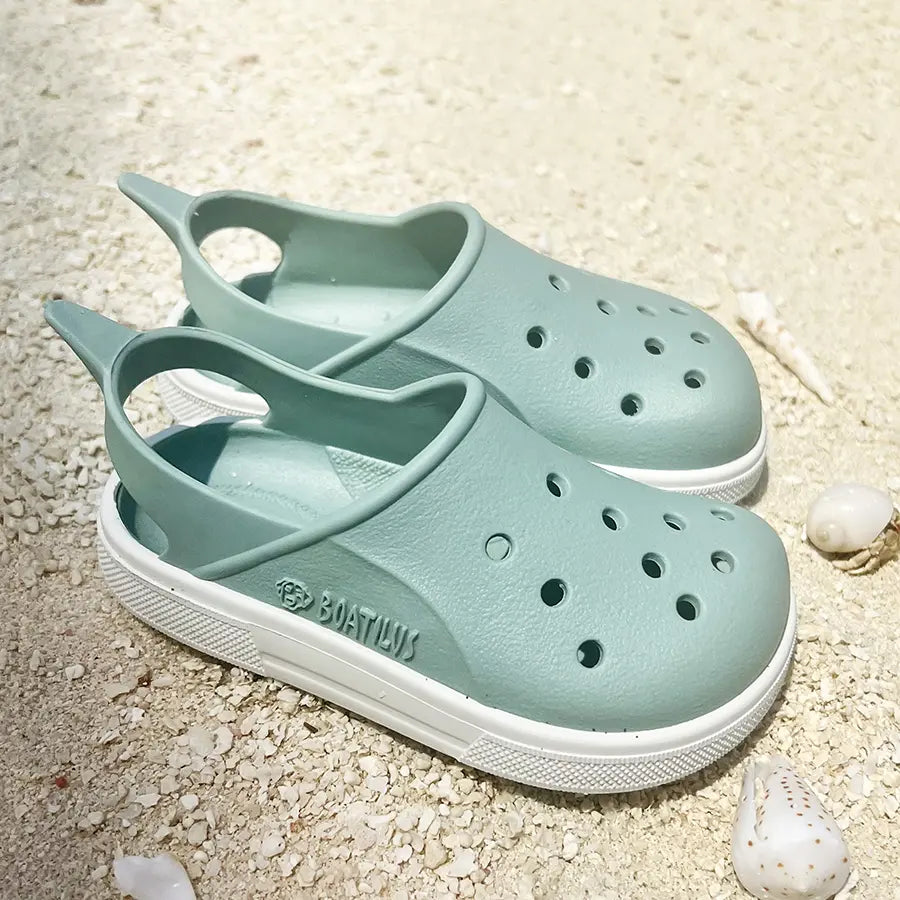 Par de sandálias respeitadoras Boatilus Cloggy Verde Pastel e Branco sobre a areia da praia.