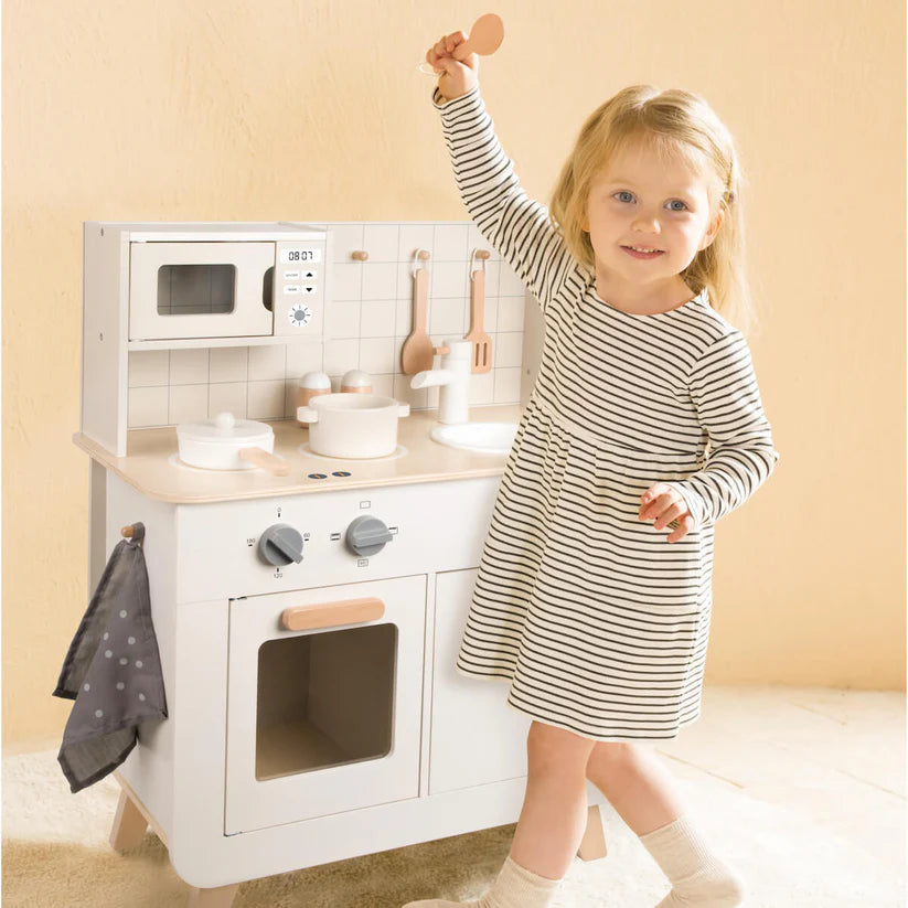 Criança loira a brincar feliz com a cozinha de madeira Classic World, segurando um utensílio de cozinha.