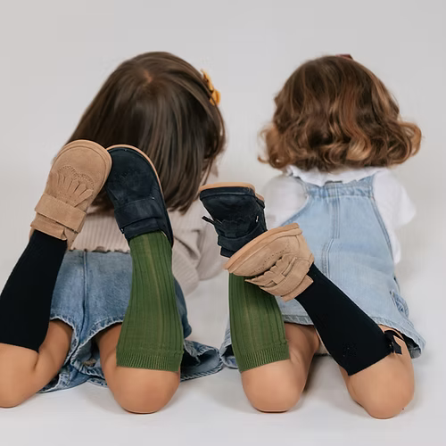 Duas crianças de costas, deitadas, a usar sapatilhas Rutis - Carneiras PiuPiu (uma em areia, outra em azul-escuro) e meias coloridas.