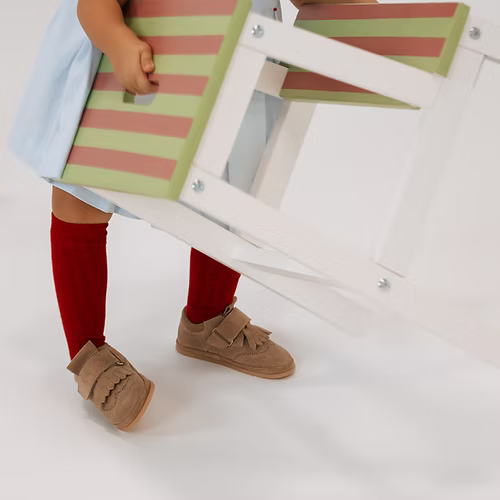 Criança em pé, a segurar um objeto, usando sapatilhas Rutis - Carneiras PiuPiu Areia e meias vermelhas, mostrando a flexibilidade do calçado.