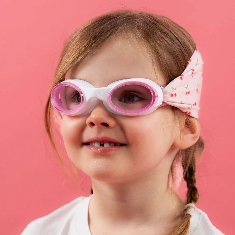 Close-up de uma menina sorridente a usar os óculos Waterplops Cerejas, exibindo a banda elástica rosa com padrão de cerejas e as lentes cor-de-rosa.