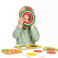 Bebé loiro a segurar uma peça de melancia do puzzle "My First Puzzle: Food" da Banana Panda à frente do rosto, num momento de diversão.