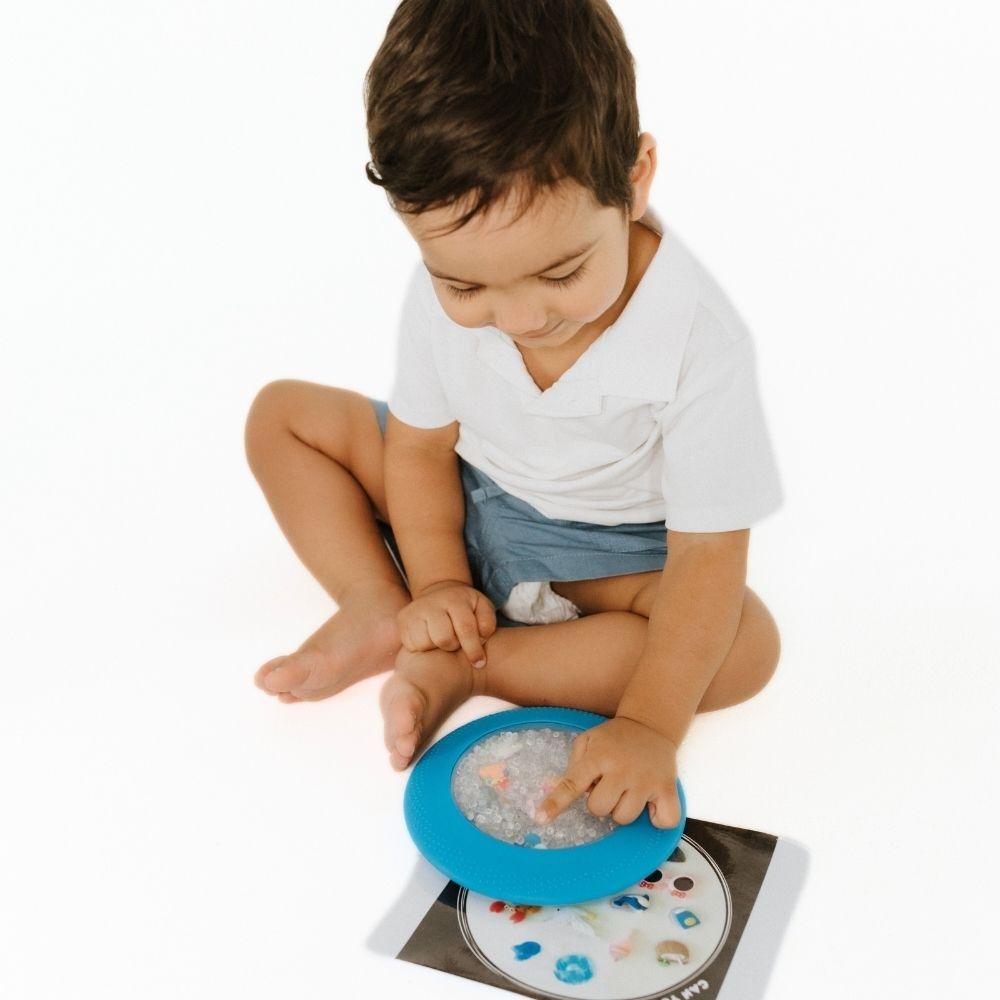 Criança sentada a interagir com a Jellystone Peekaboo Bag azul e folha de atividades.