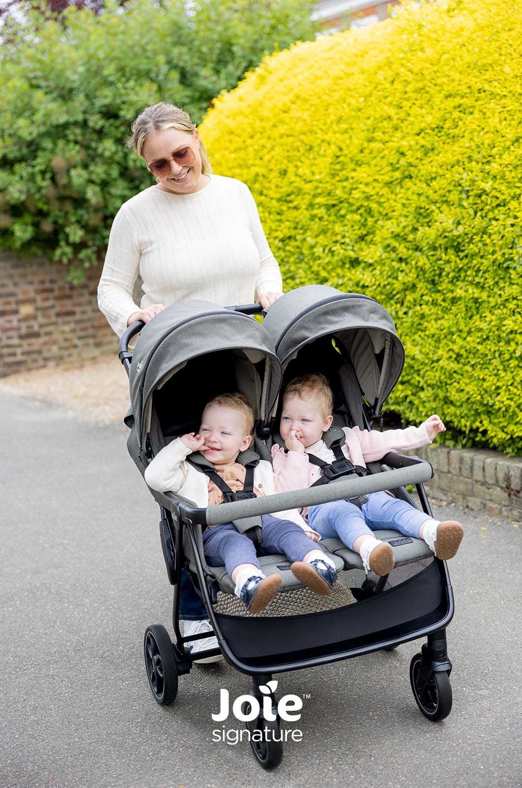 Mãe sorridente a passear com o carrinho duplo Joie Estrella Signature, com dois bebés felizes sentados.