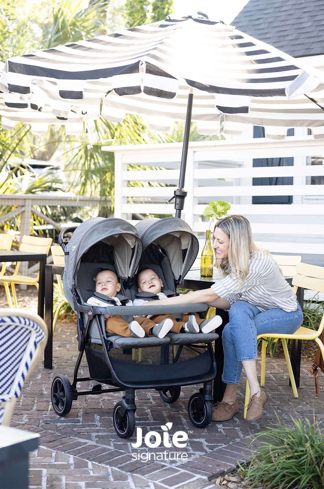 Mãe com dois bebés num carrinho duplo Joie Estrella Signature, sentados à mesa de um café exterior com um chapéu de sol listrado.