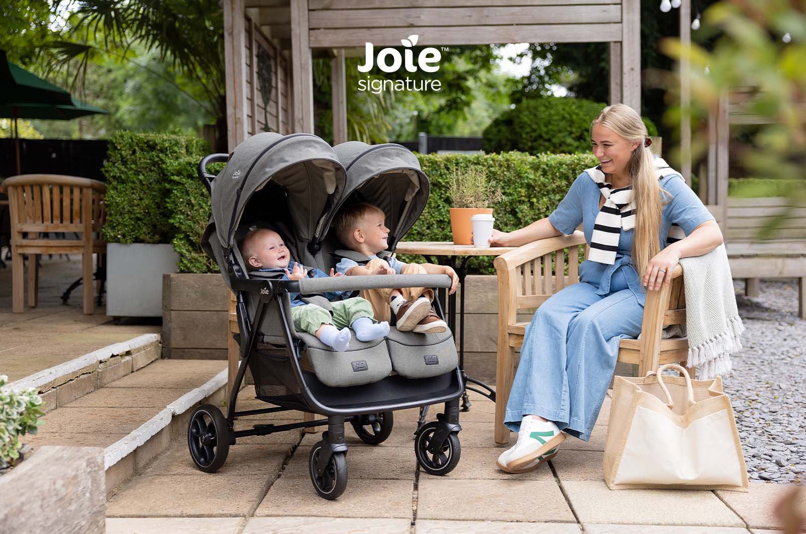 Mãe sentada numa mesa exterior com o carrinho Joie Estrella Signature e dois bebés ao lado.