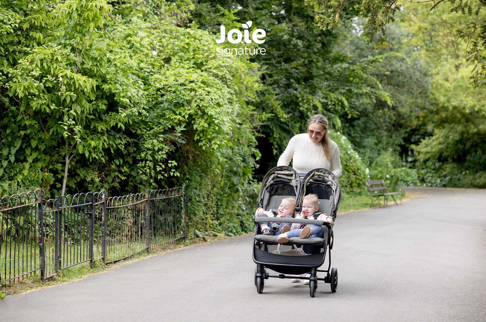 Mãe a empurrar o carrinho duplo Joie Estrella Signature com dois bebés num caminho de parque.