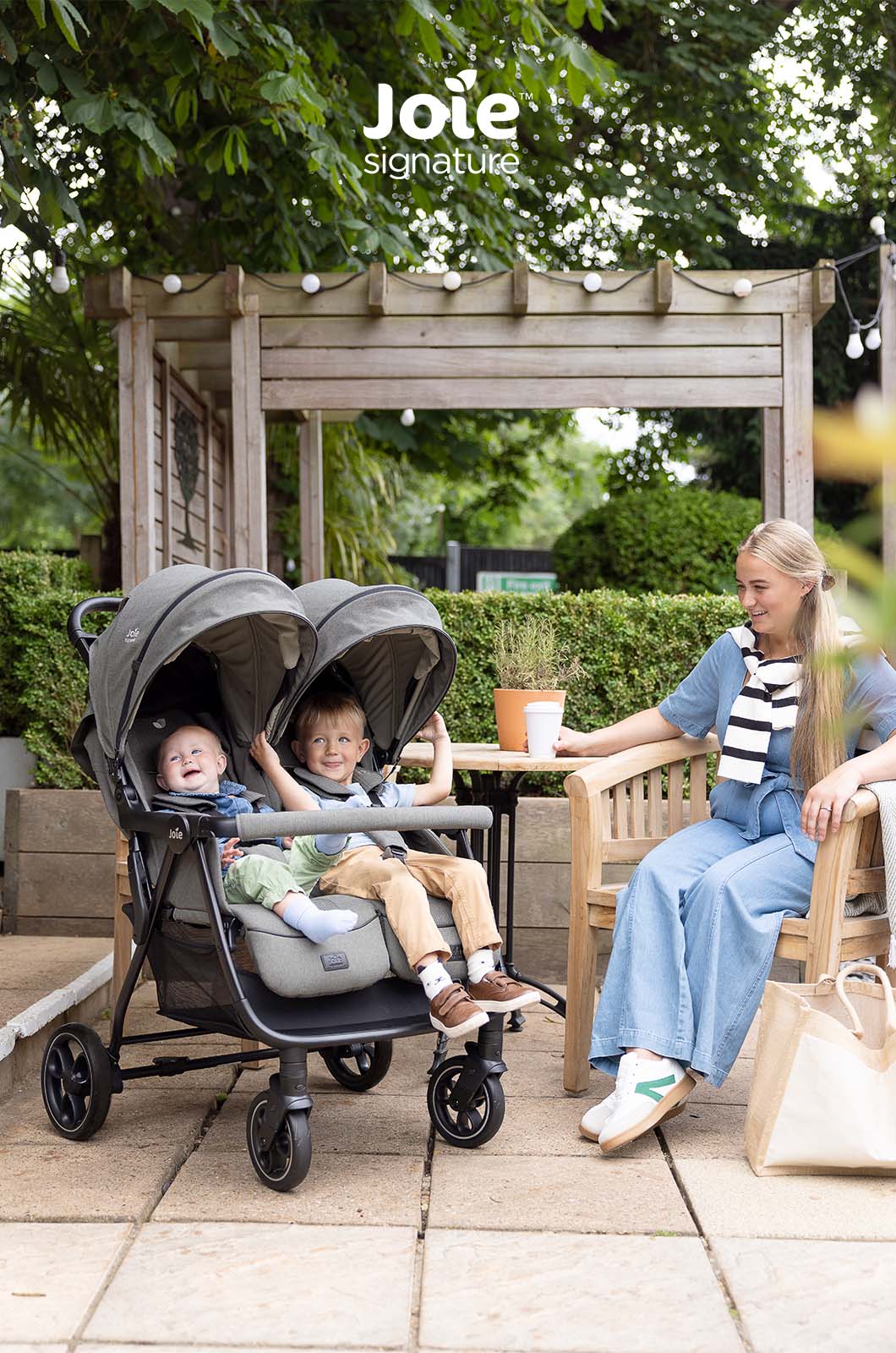 Mulher a passear com o carrinho duplo Joie Estrella Signature perto da entrada de uma casa, vista lateral.