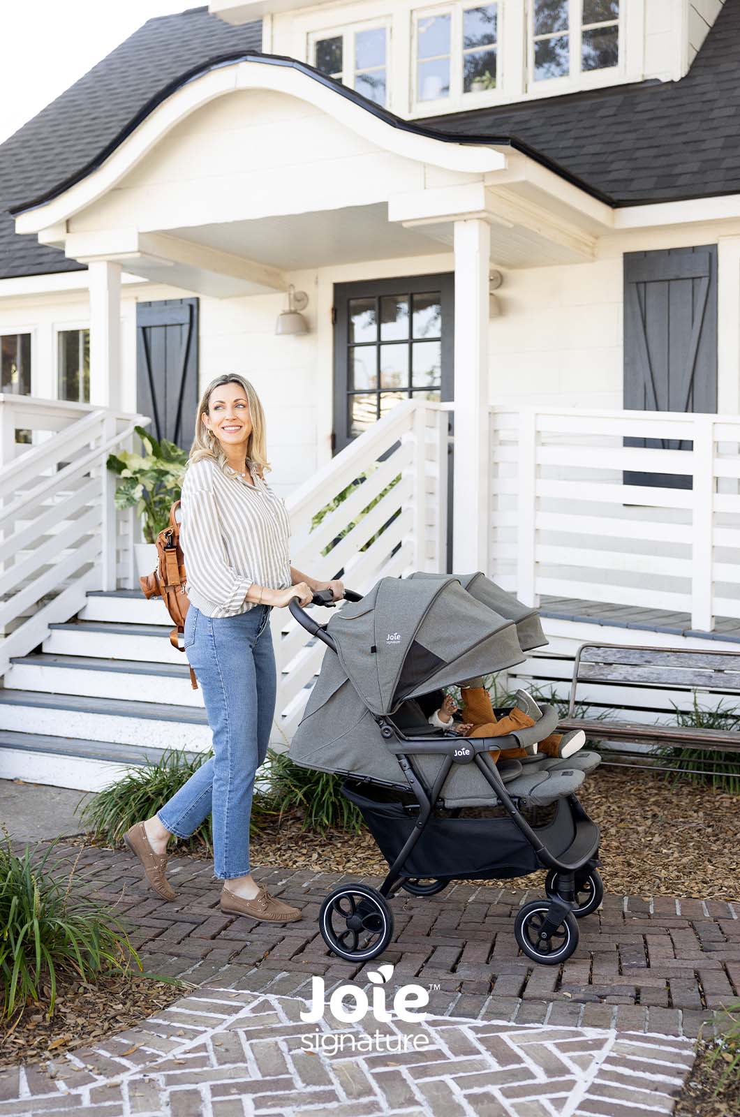 Mãe a passear com o carrinho duplo Joie Estrella Signature com um bebé perto da entrada de uma casa.