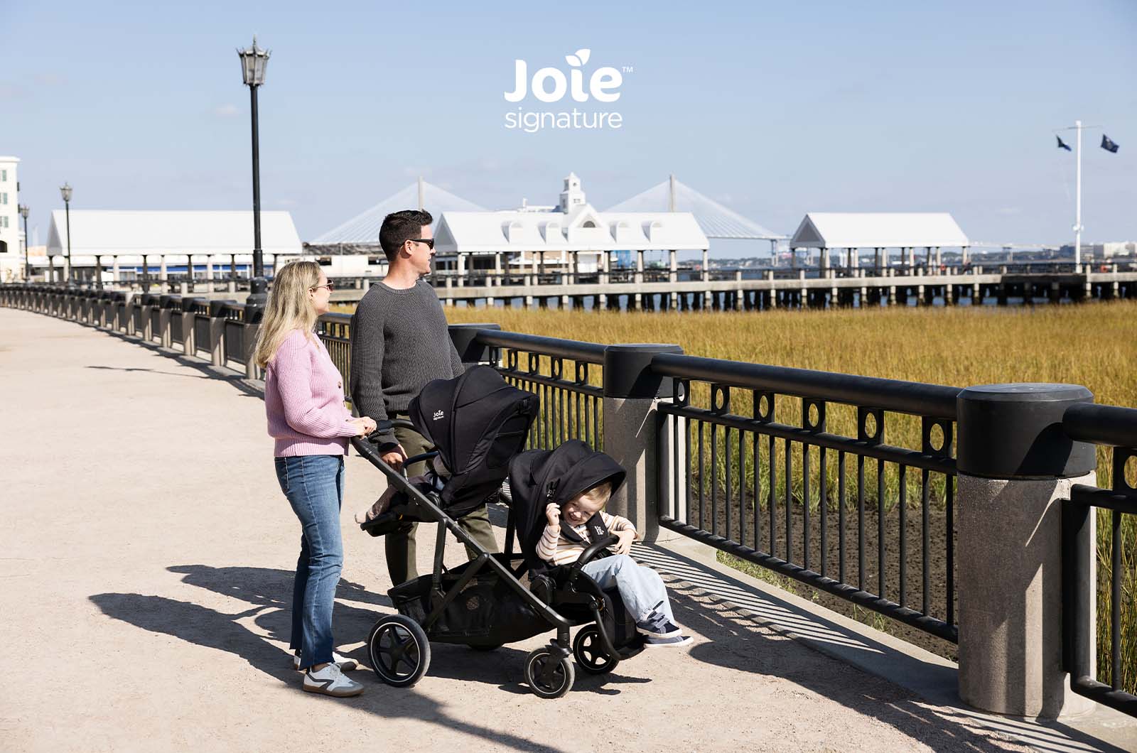 Família a passear com carrinho Joie Versiti Signature configurado para duas crianças (uma em assento, outra em alcofa) junto à água.