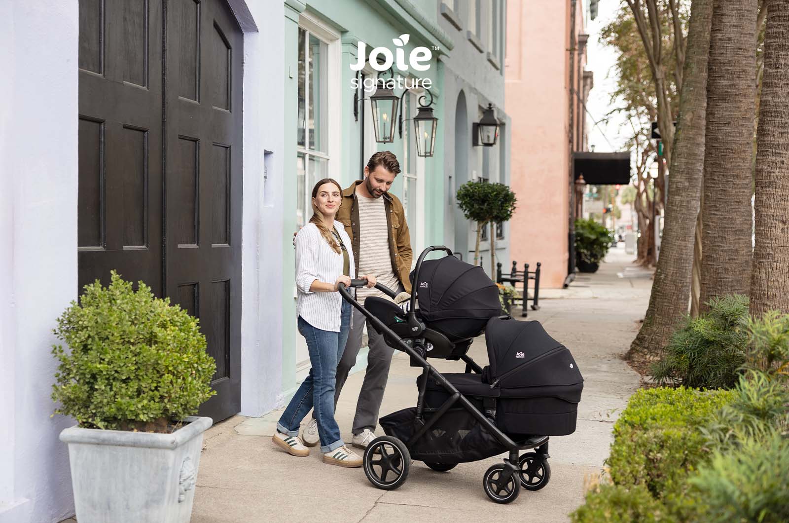 Família a passear com carrinho Joie Versiti Signature configurado para duas crianças (uma em assento, outra em alcofa) em rua.