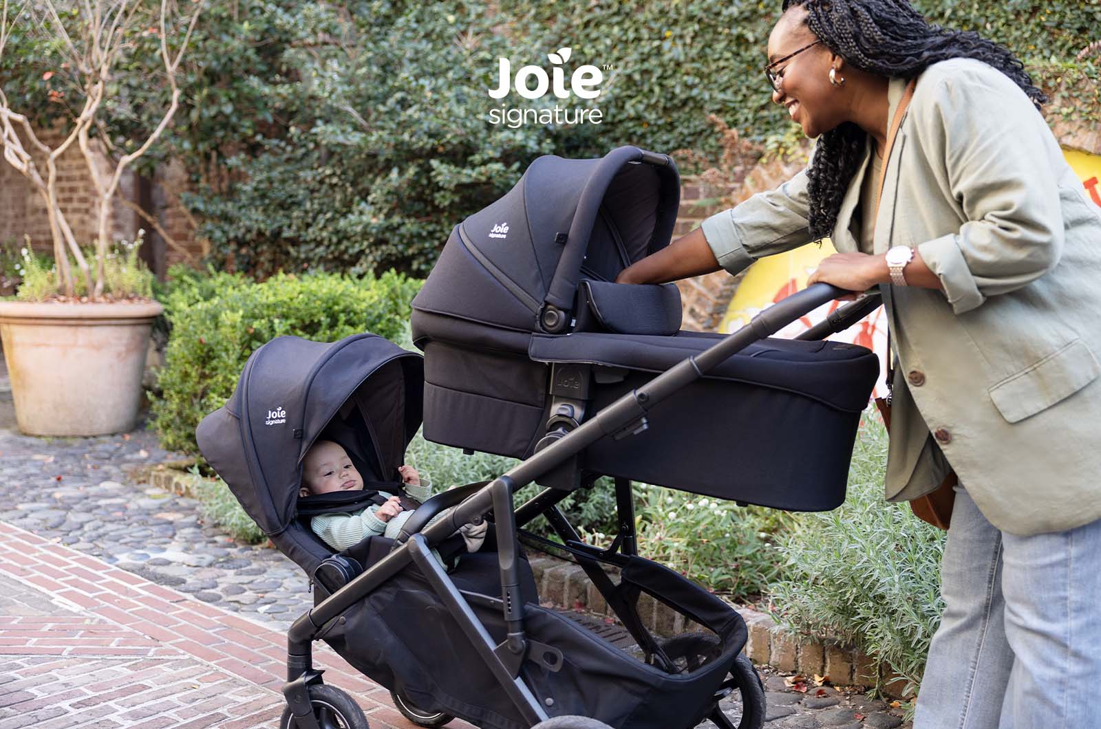 Mãe a interagir com bebé numa alcofa no carrinho Joie Versiti Signature, com segundo assento vazio ao lado.