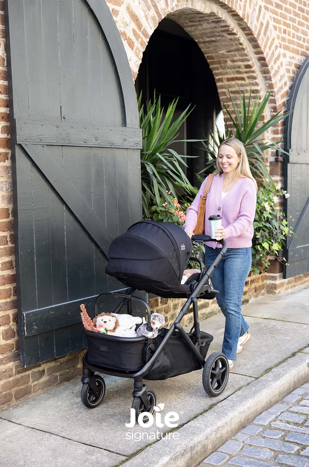 Mãe a passear com carrinho Joie Versiti Signature configurado para uma criança, com cesto cheio.