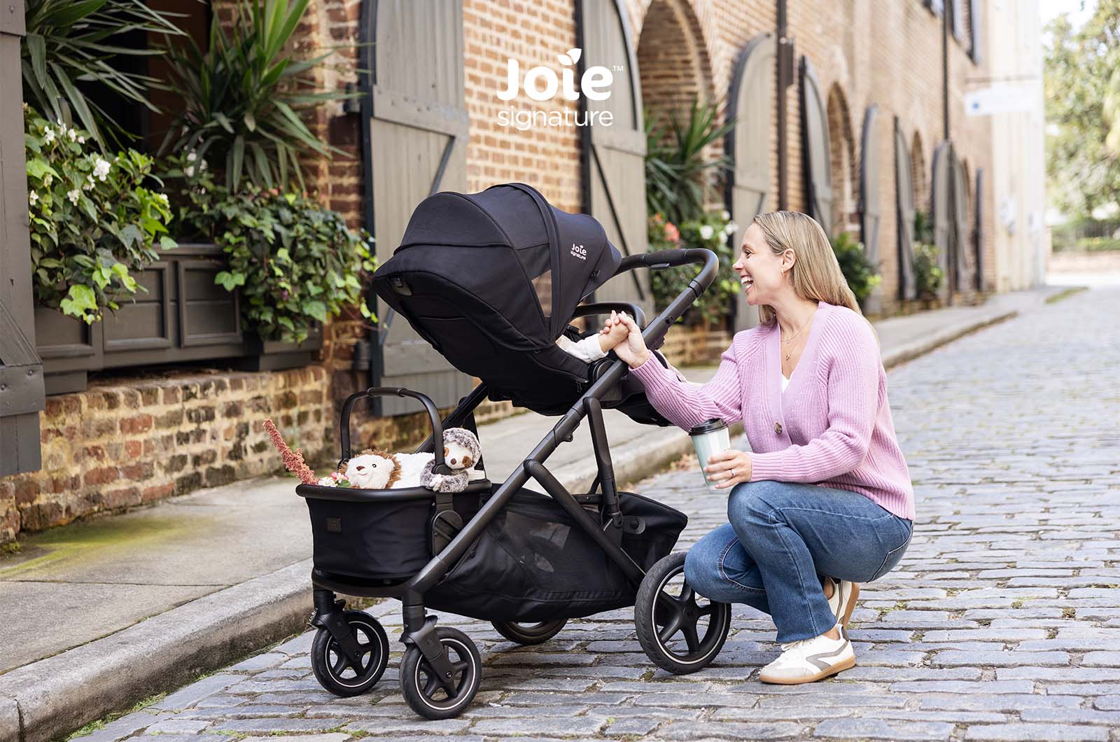 Mãe agachada a interagir com bebé no carrinho Joie Versiti Signature, cesto cheio.