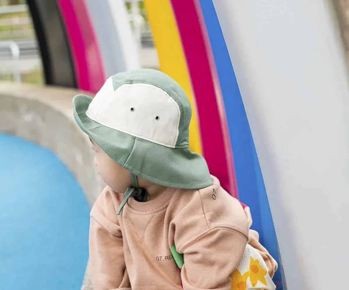 Bebé usando o chapéu Panamá Kietla verde e natural, num parque infantil colorido.  Proteção solar e estilo para crianças.