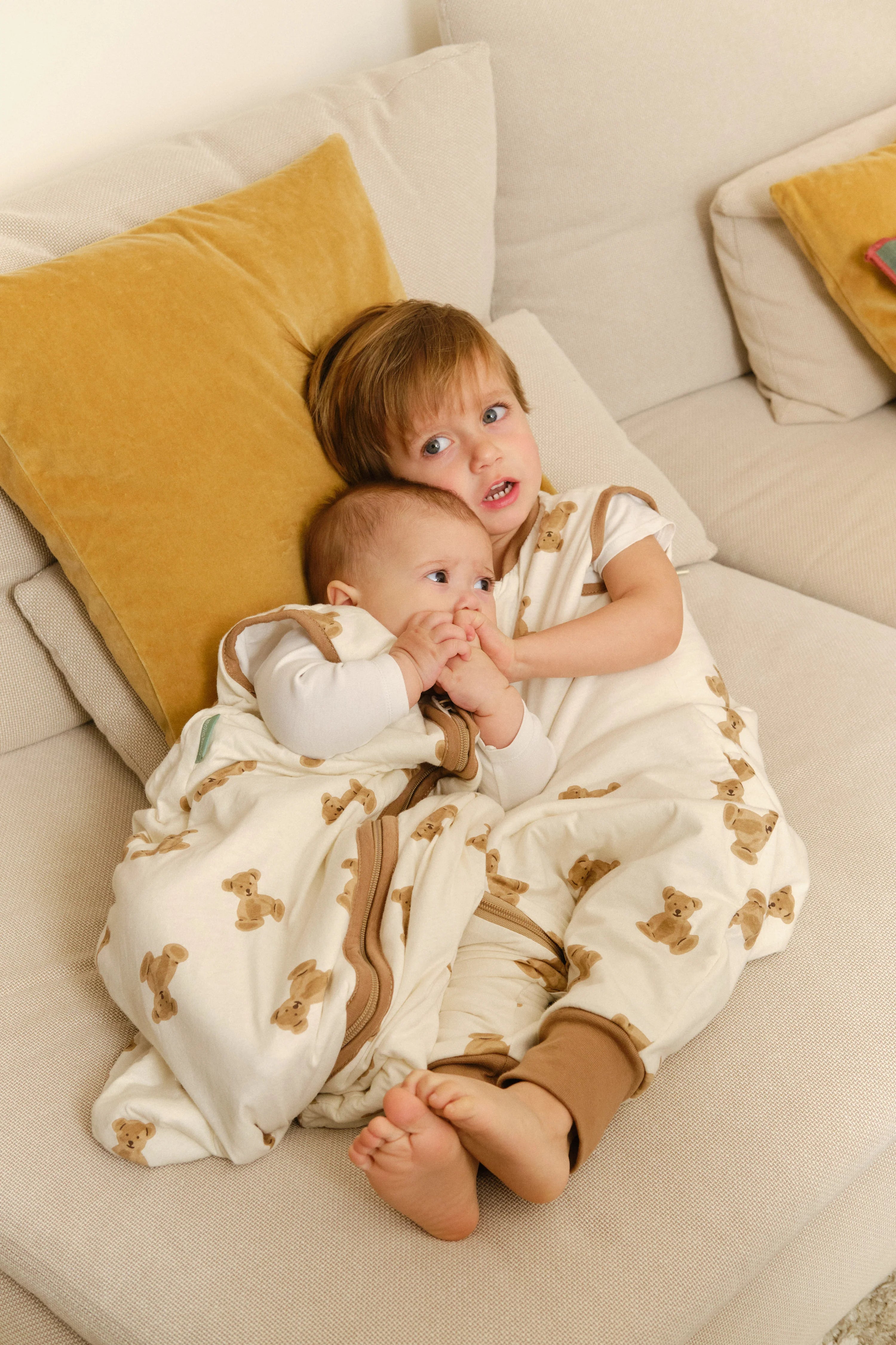 Bebé aconchegado em saco de dormir MolisandCo TOG 1.0 Teddy creme, abraçado por criança no sofá. Conforto e segurança.