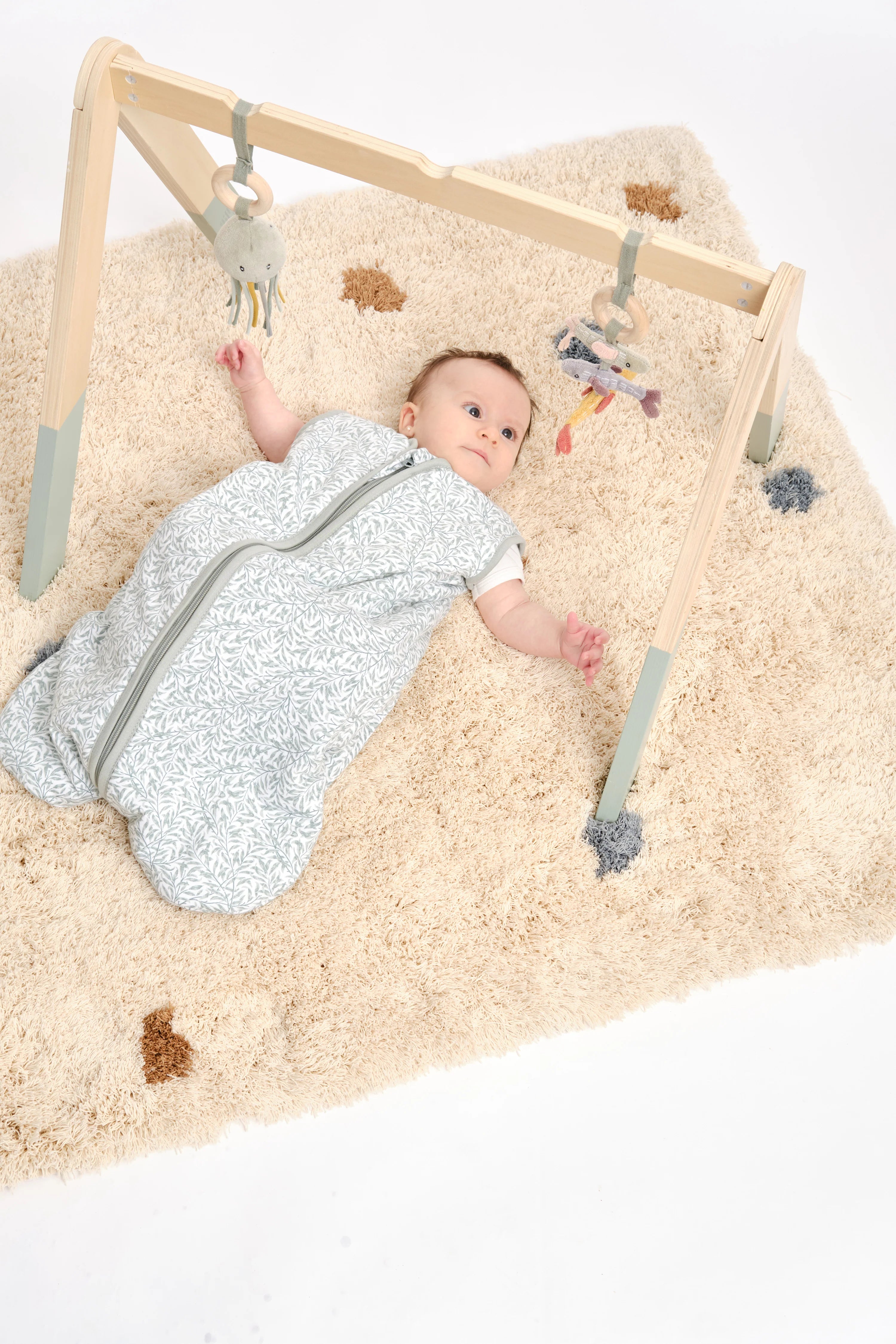 Bebé no saco de dormir MolisandCo TOG 1.0 Ines II, deitado numa manta felpuda sob um ginásio de atividades em madeira.