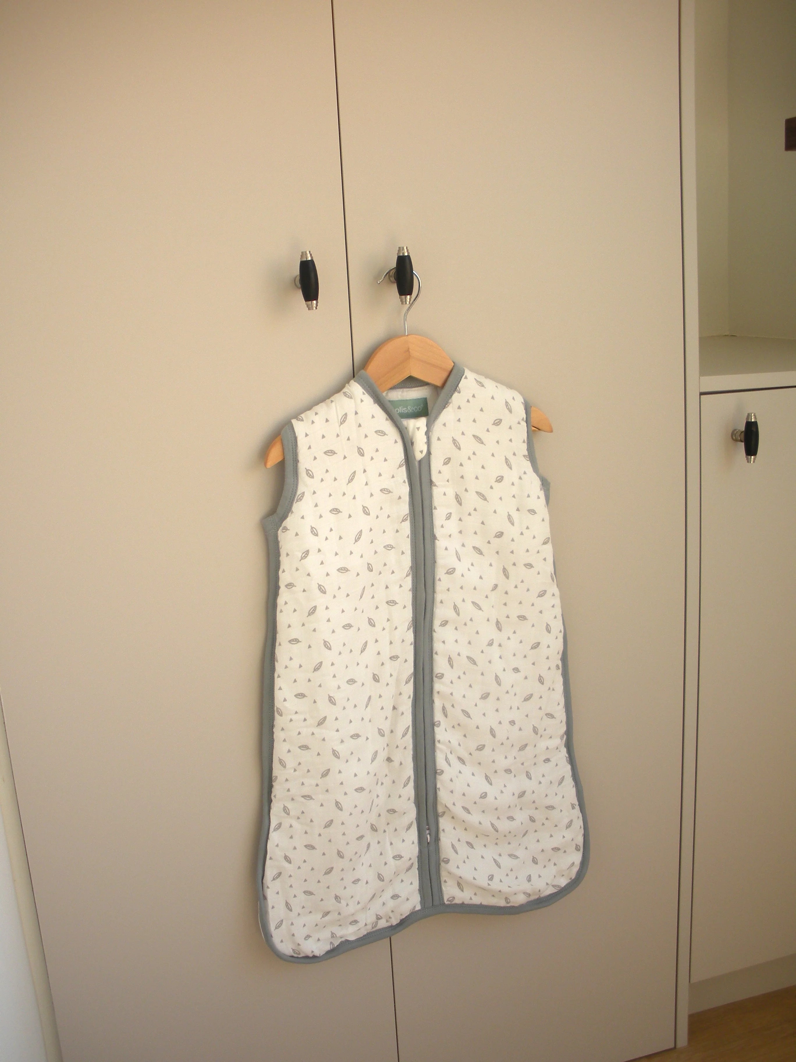 Saco de dormir MolisandCo TOG 1.0 de musselina, padrão folhas, pendurado num cabide de madeira em armário.