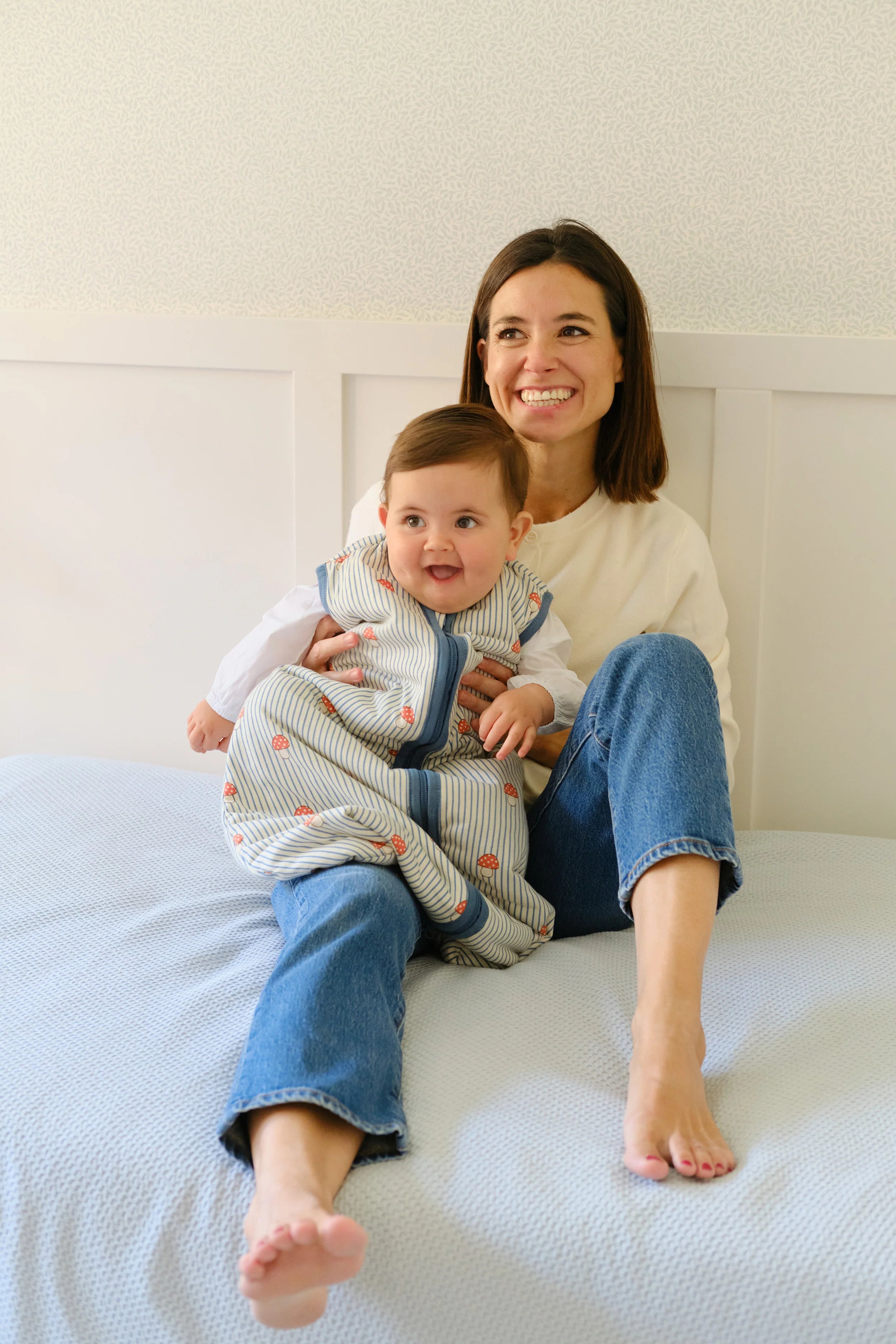 Mãe sorridente segura bebé feliz em saco de dormir MolisandCo TOG 1.0 'Mushrooms' na cama.