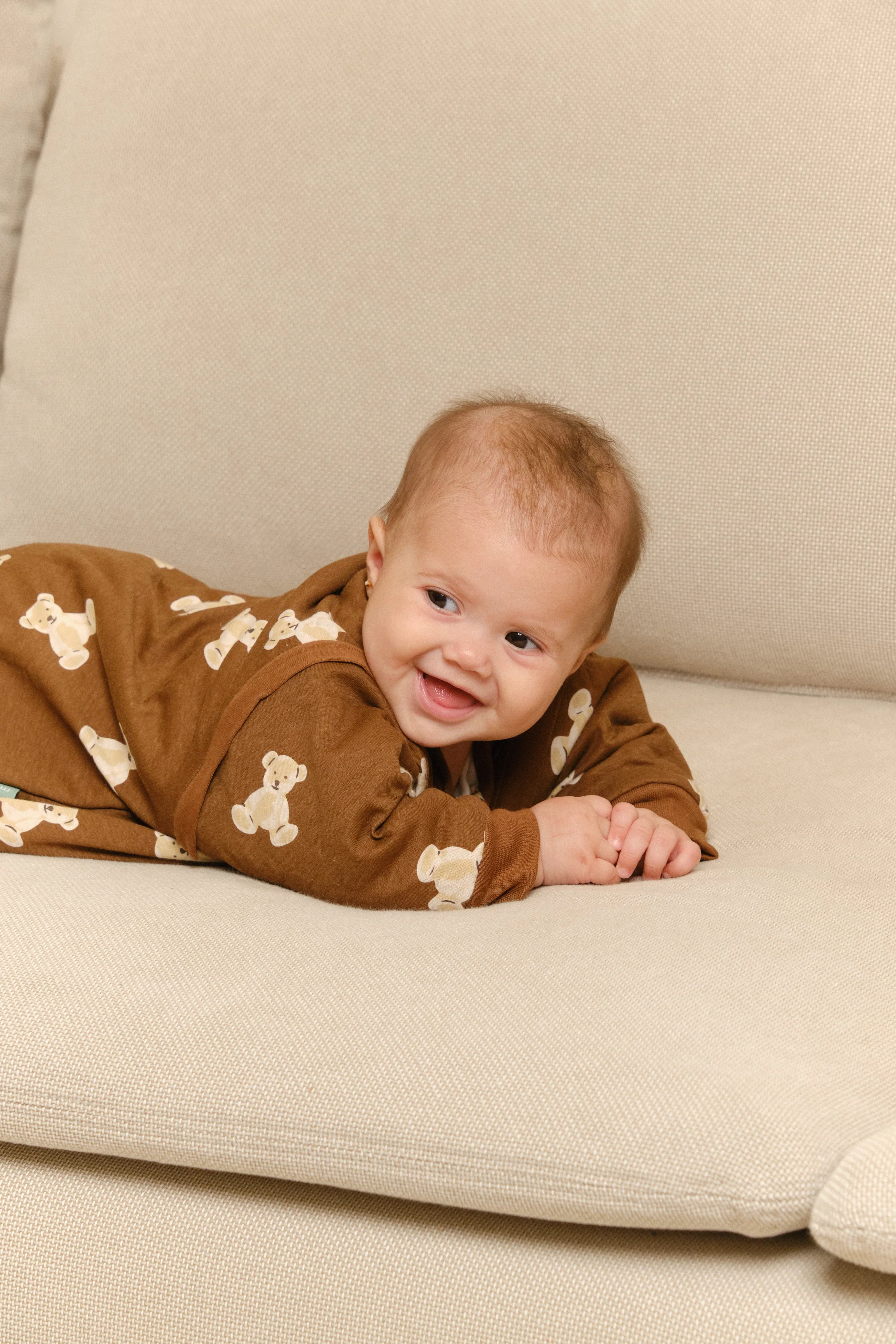 Um bebé sorridente deitado de bruços num sofá, usando o saco de dormir MolisandCo TOG 1.0 Teddy Mocca castanho.