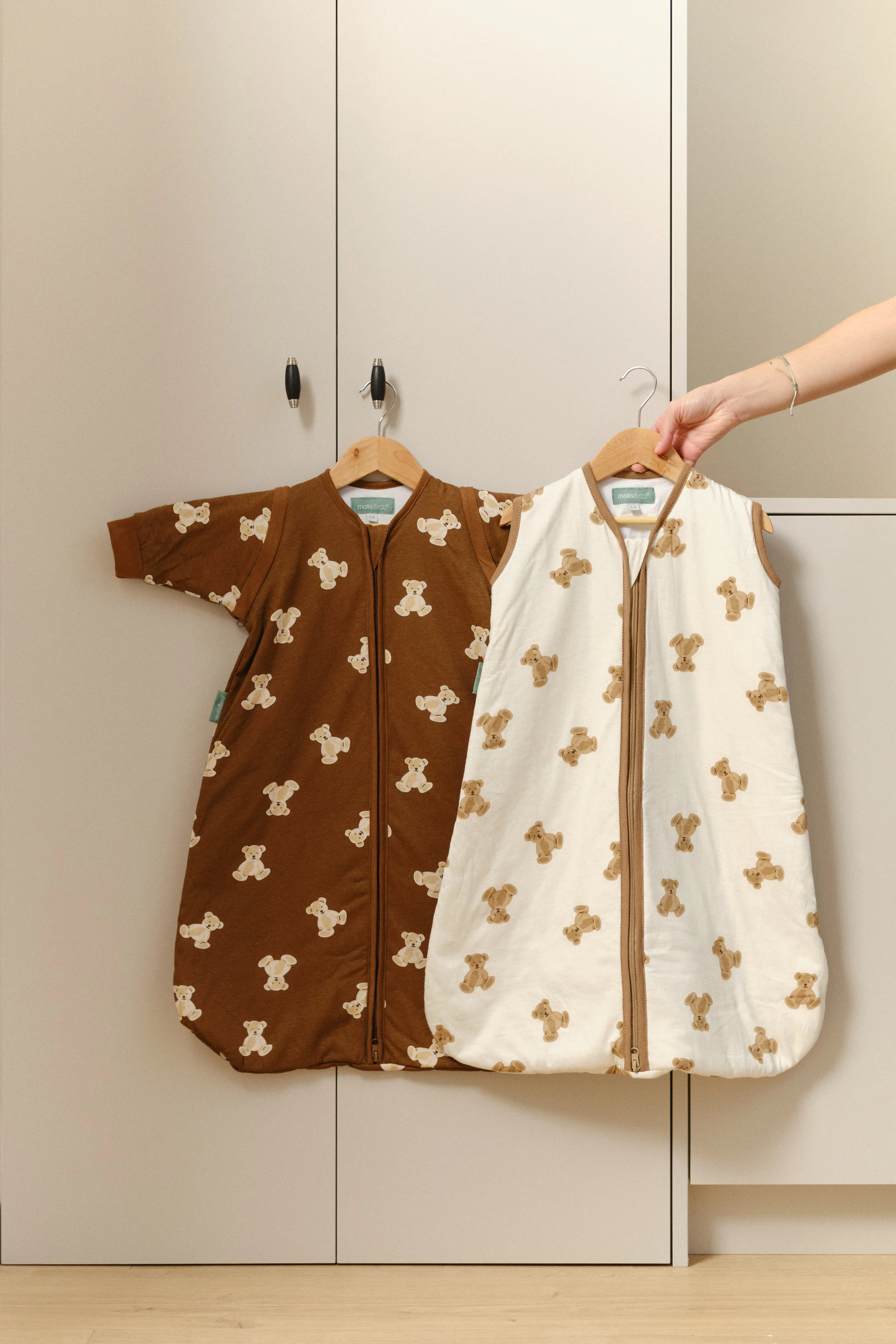 Dois sacos de dormir MolisandCo TOG 1.0 Teddy Mocca, um castanho e um branco, pendurados num armário, com uma mão a segurar o branco.