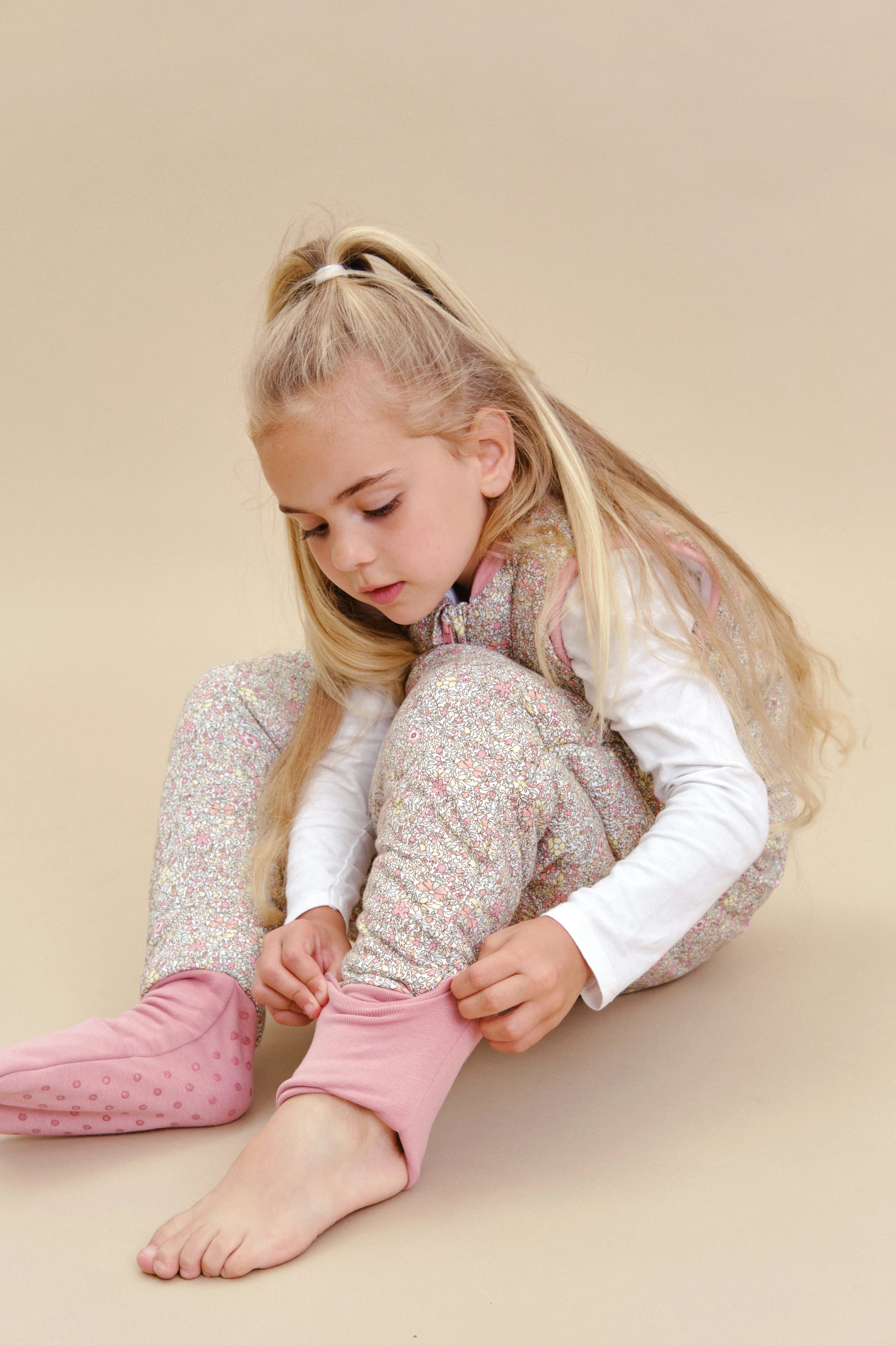 Criança sentada a ajustar punho do saco de dormir MolisandCo TOG 2.5 Pastel Floral, com meias removíveis.