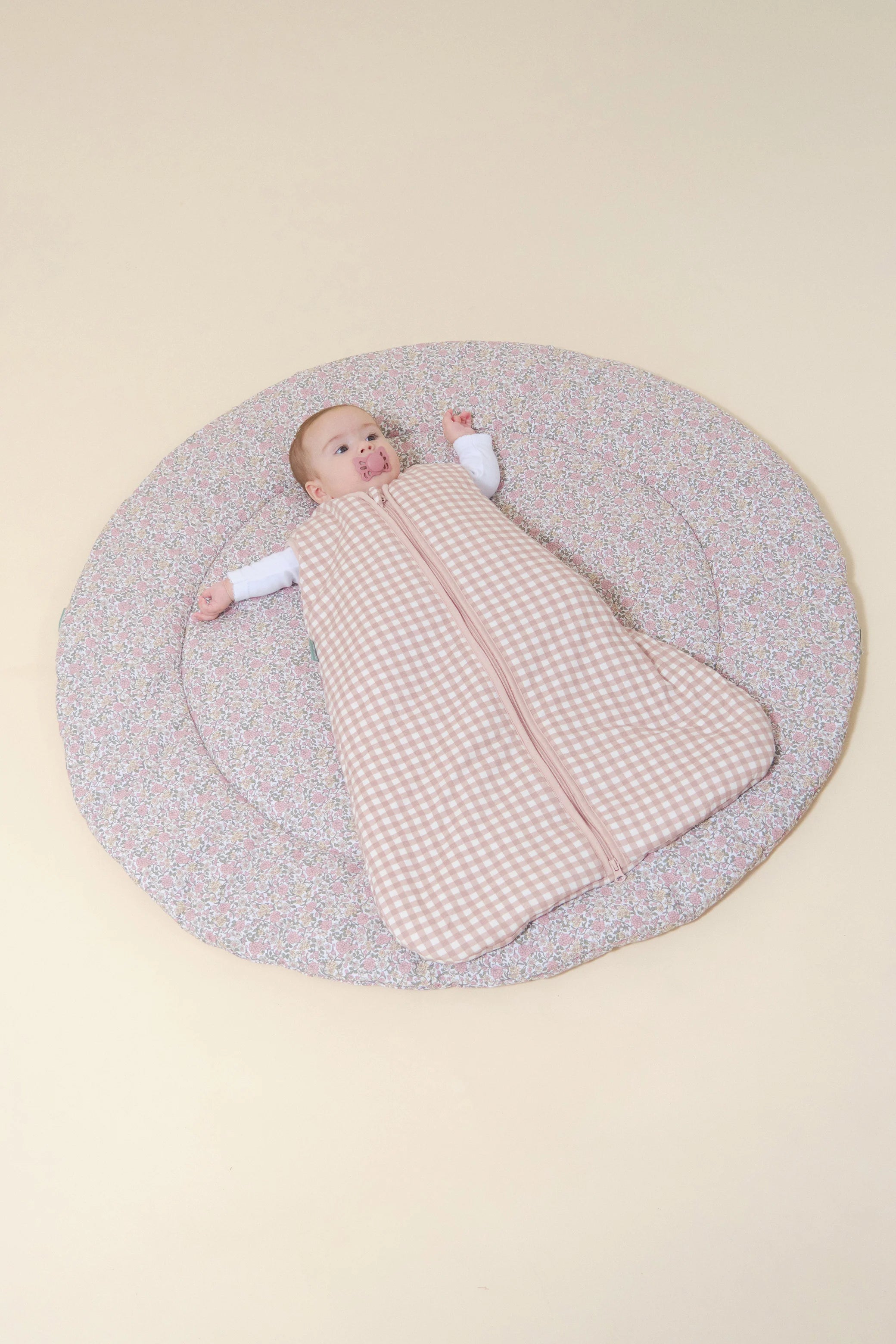 Bebé deitado a usar saco de dormir MolisandCo TOG 2.5 Dusty Rose xadrez, sem pés, em tapete florido.