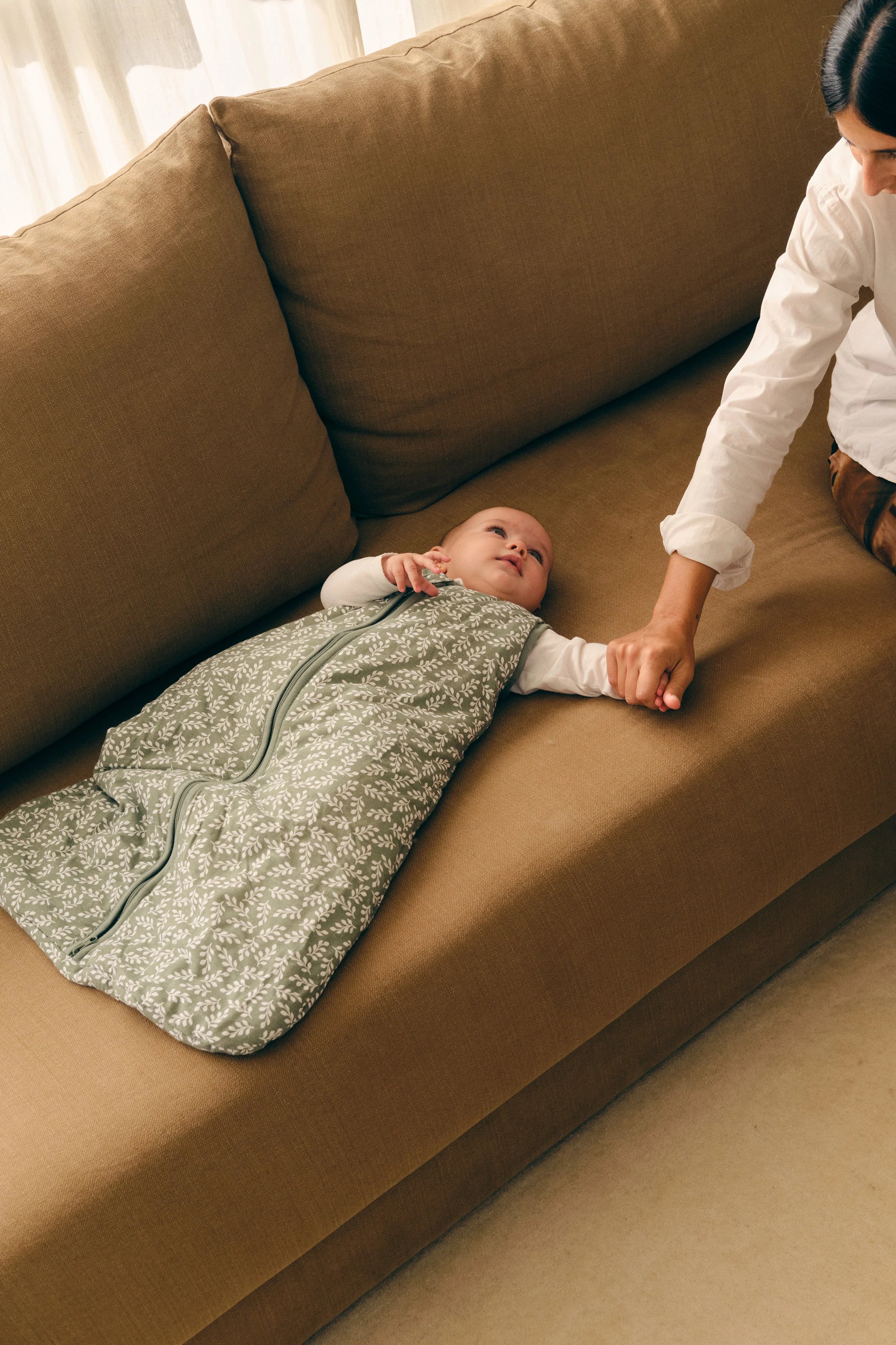 Bebé feliz no saco de dormir MolisandCo TOG 2.5 Green Garden, deitado no sofá, com a mão da mãe.