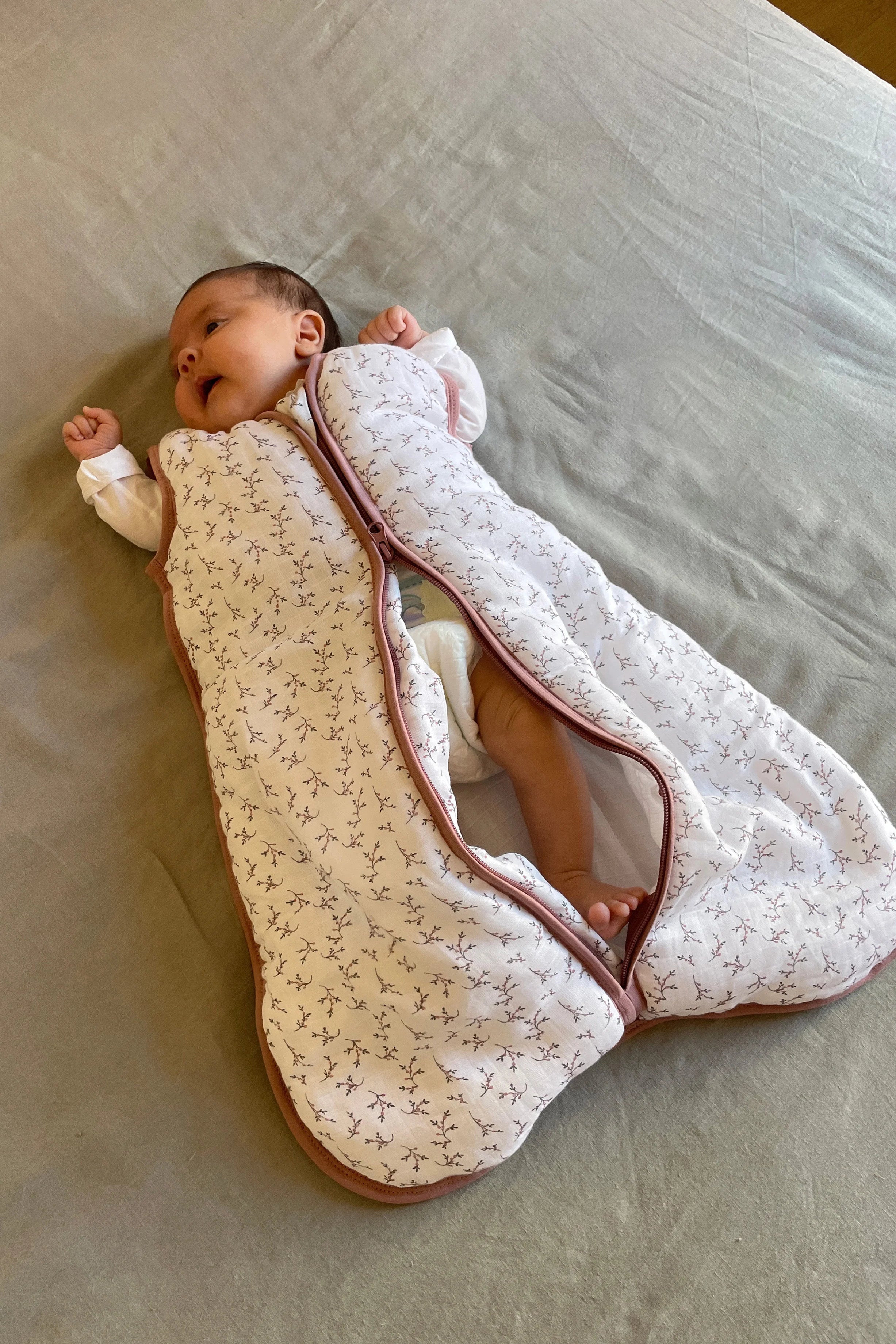 Bebé deitado na cama, com o saco de dormir MolisandCo TOG 2.5 rosa floral parcialmente aberto, mostrando as pernas.