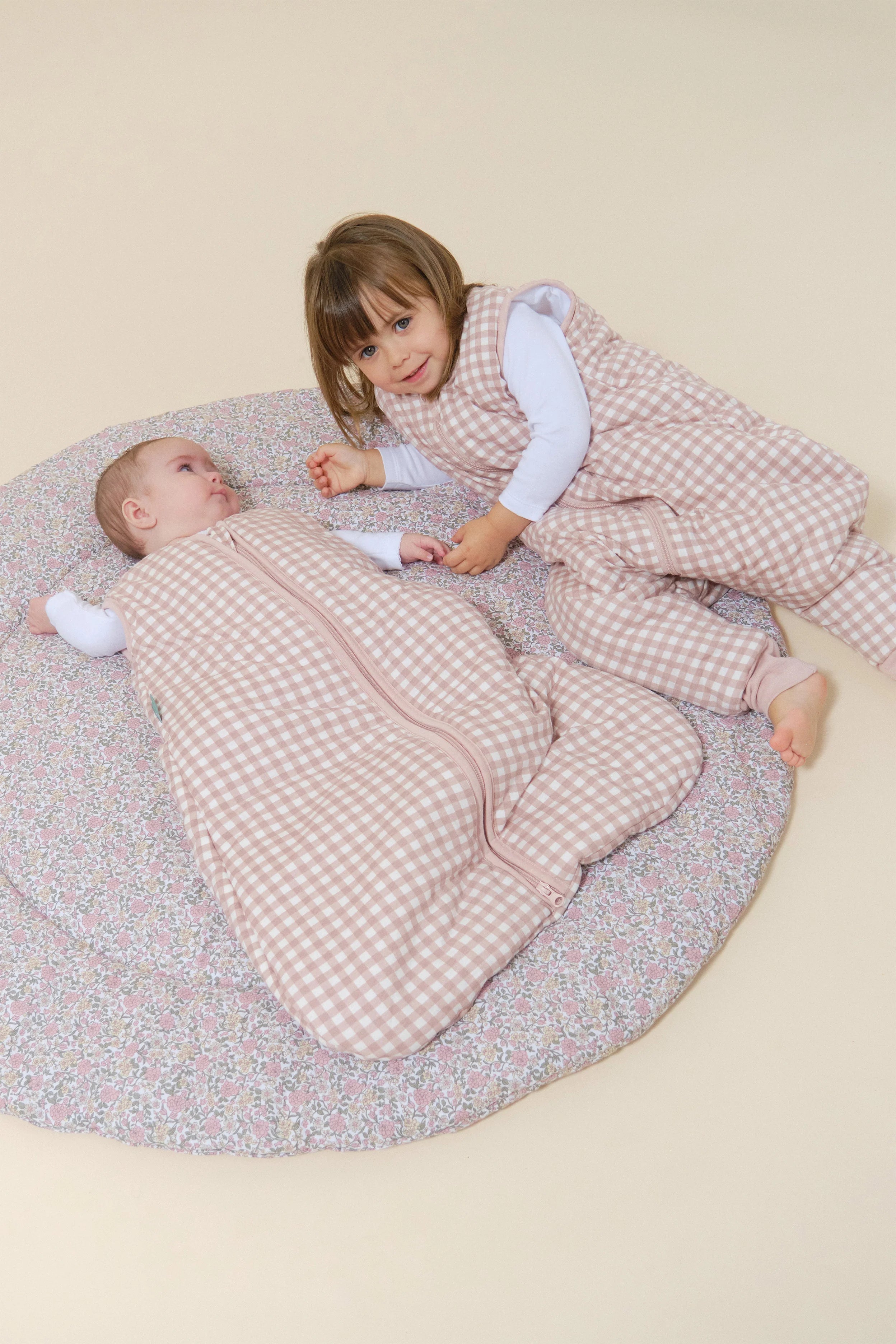 Duas crianças, bebé e criança mais velha, deitadas em tapete, com sacos de dormir MolisandCo TOG 2.5 com pés rosa xadrez.