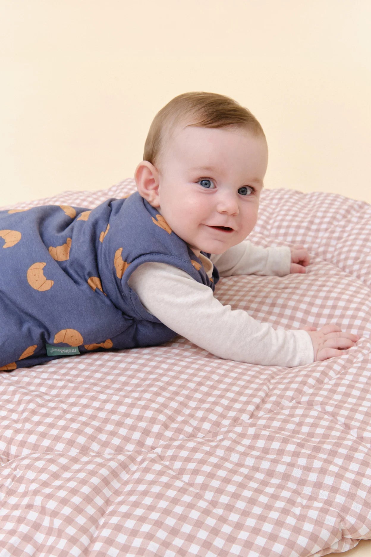 Bebé sorridente deitado de bruços, com saco de dormir MolisandCo TOG 2.5 azul de croissants, mostrando o padrão.