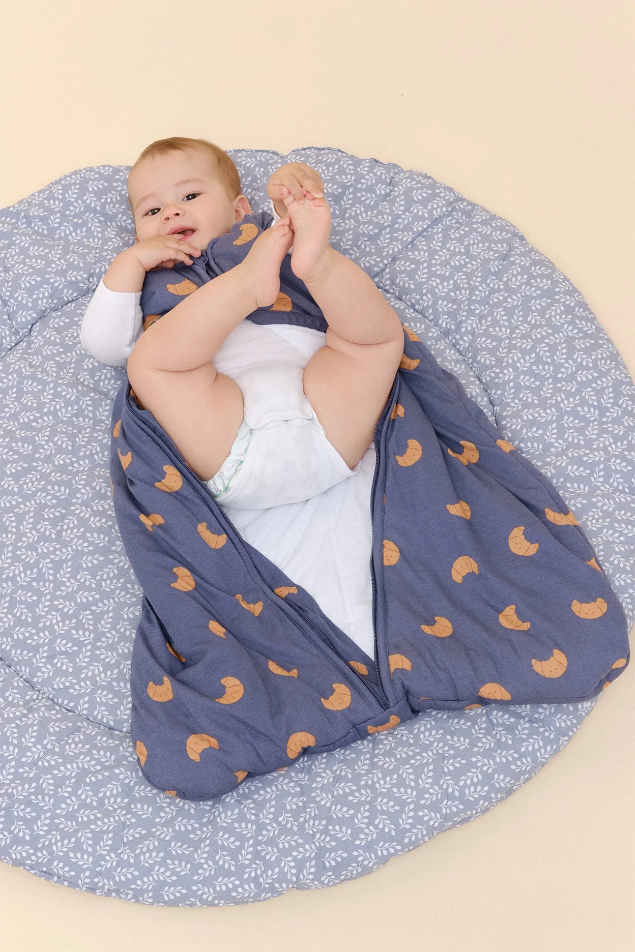 Bebé deitado de costas, com saco de dormir MolisandCo TOG 2.5 azul de croissants aberto, mostrando as pernas.