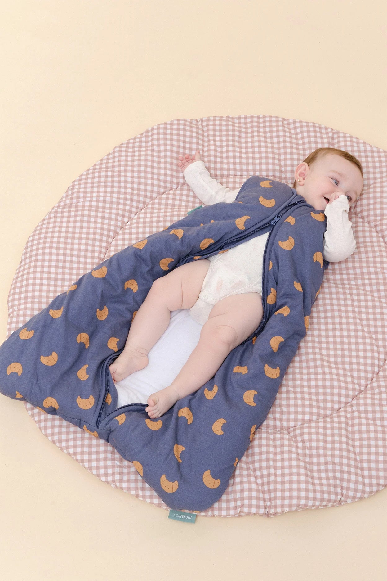 Bebé deitado de costas, relaxado no saco de dormir MolisandCo TOG 2.5 azul de croissants, parcialmente aberto.