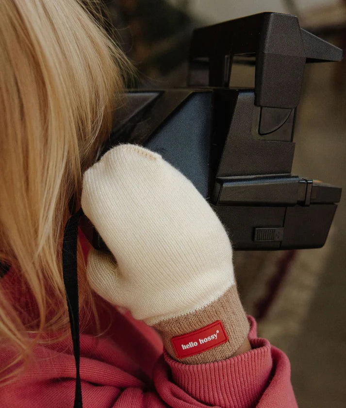 Criança loira a usar as luvas Hello Hossy Hotty Sandy creme, a segurar uma máquina fotográfica instantânea vintage.