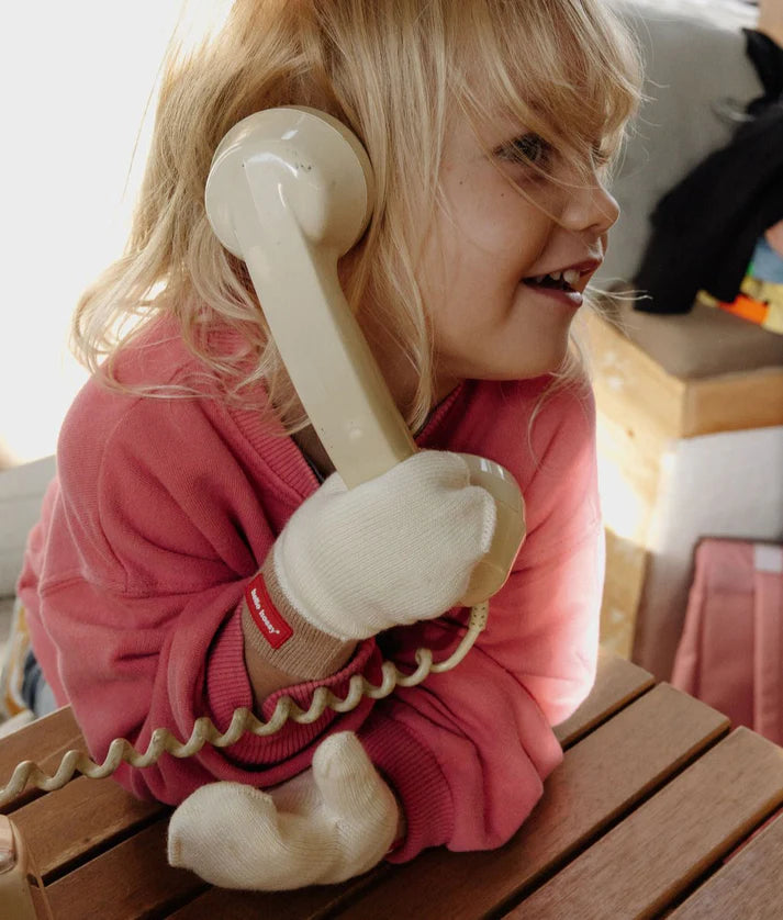 Criança loira sorridente a segurar um telefone antigo, usando as luvas Hello Hossy Hotty Sandy creme com punho castanho e etiqueta vermelha.